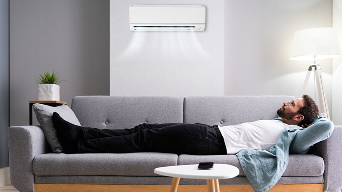 Pessoa deitada num sofá cinzento numa sala moderna; ar condicionado na parede acima, mesa de centro branca à frente e candeeiro de pé junto à parede.